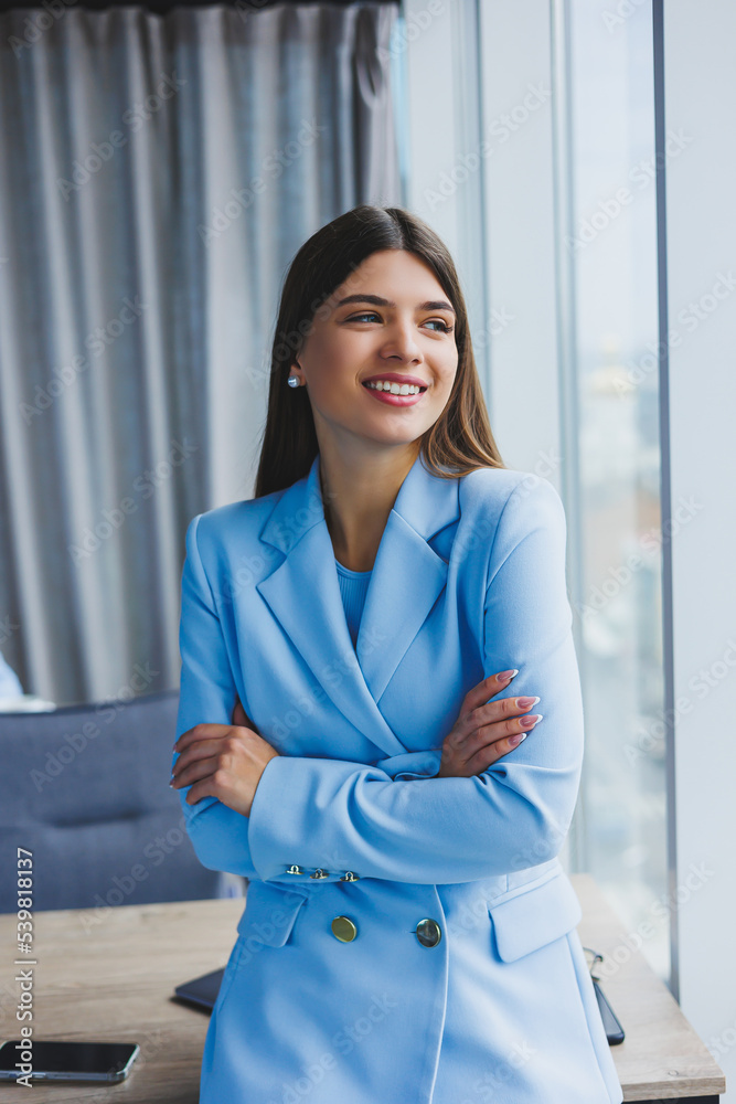 Portrait of a pretty brunette in a blue jacket in the office near the panoramic window with a view of the city. Happy business woman in the office Portrait of a pretty brunette in a blue jacket in the office near the panoramic window with a view of the city. Happy business woman in the office