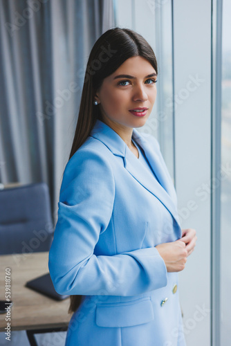 Free Download Portrait of a pretty brunette in a blue jacket in the office near the panoramic window with a view of the city. Happy business woman in the office - GFXLoad