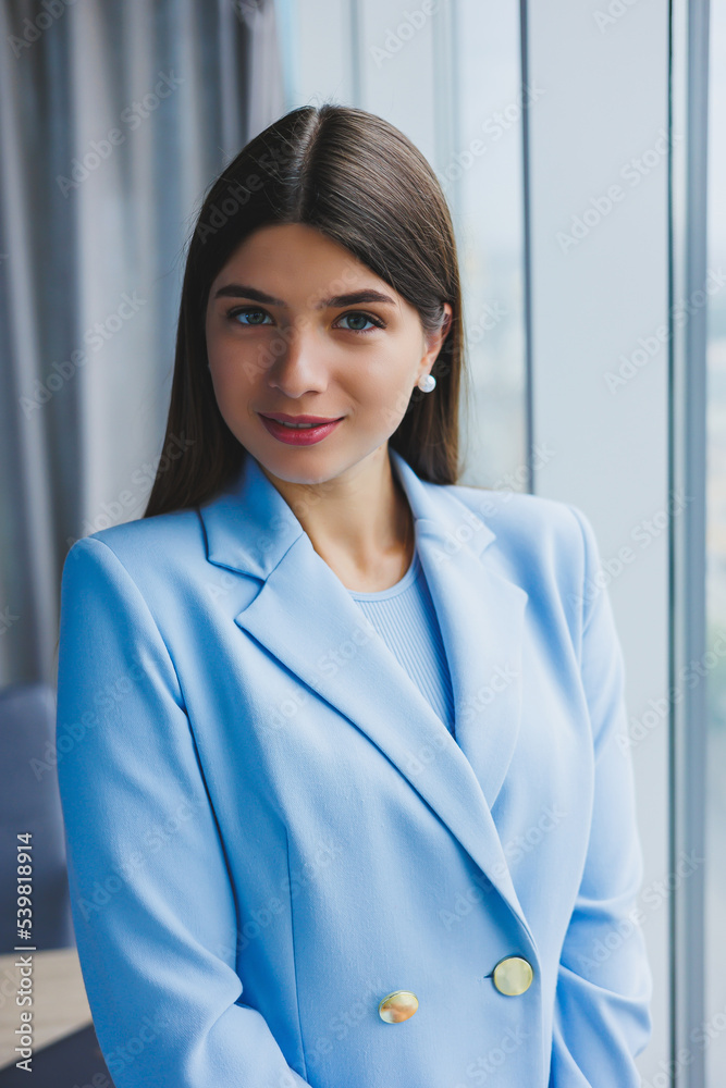 Portrait of a pretty brunette in a blue jacket in the office near the panoramic window with a view of the city. Happy business woman in the office Portrait of a pretty brunette in a blue jacket in the office near the panoramic window with a view of the city. Happy business woman in the office