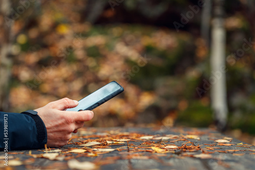 A man uses a smartphone in his hands in the autumn forest at an old wooden table. The background is blurry.