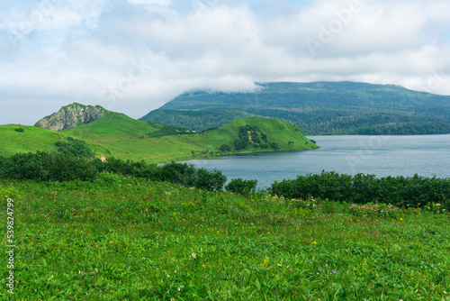 natural landscape of Kunashir island with grassy hills, volcanic rocks, volcano in the clouds and a valley with a lagoon