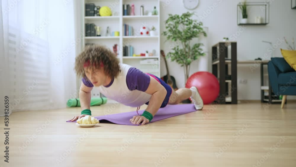 Amusing young man doing push-ups at home using croissant as a motivation Amusing young man doing push-ups at home using croissant as a motivation