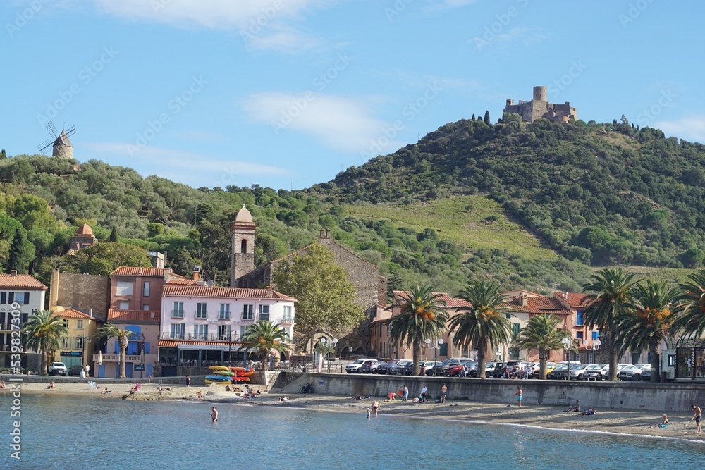 Collioure, France - October 2022; View across bay featuring people in water and on beach against backdrop of colourful buildings, and hill featuring windmill and Fort Saint Elme 