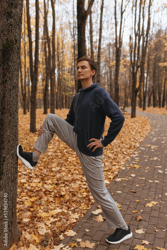 Fit male athlete in activewear leaning on tree and stretching legs while warming up during outdoor workout in autumn park 