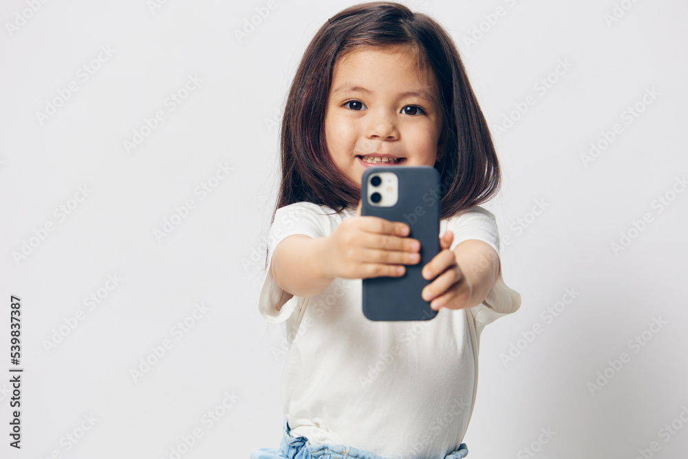 a cute little preschool girl stands on a white background in a white T-shirt and shows her phone to the camera, smiling happily. The theme of children's happiness a cute little preschool girl stands on a white background in a white T-shirt and shows her phone to the camera, smiling happily. The theme of children's happiness