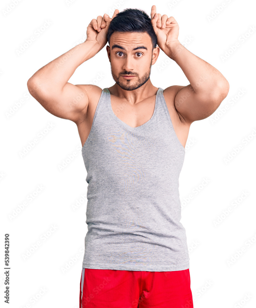 Young handsome man wearing swimwear and sleeveless t-shirt doing funny gesture with finger over head as bull horns Young handsome man wearing swimwear and sleeveless t-shirt doing funny gesture with finger over head as bull horns
