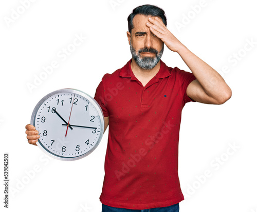 Middle aged man with beard holding big clock stressed and frustrated with hand on head, surprised and angry face