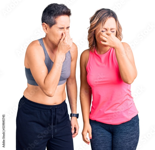 Couple of women wearing sportswear smelling something stinky and disgusting, intolerable smell, holding breath with fingers on nose. bad smell