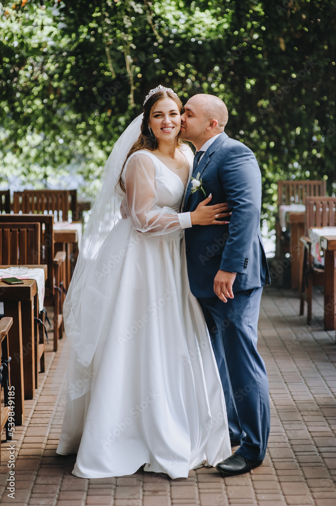 Stylish adult groom in a blue suit and a beautiful young bride hugging while standing in the park. Wedding photography of the newlyweds, portrait.