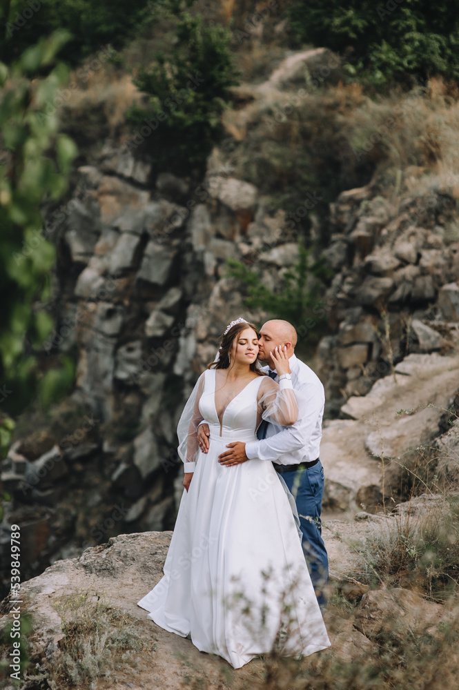 An adult groom and a beautiful, sweet bride in a white dress are embracing in the open air, against the backdrop of rocks, mountain cliffs. Wedding photography, portrait.
