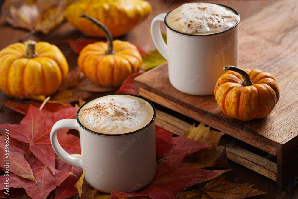 Two white cups with black coffee and white milk foam and cinnamon spice in a cozy autumn setting with red, yellow and orange leaves, hokkaido pumpkins, on wooden background Two white cups with black coffee and white milk foam and cinnamon spice in a cozy autumn setting with red, yellow and orange leaves, hokkaido pumpkins, on wooden background