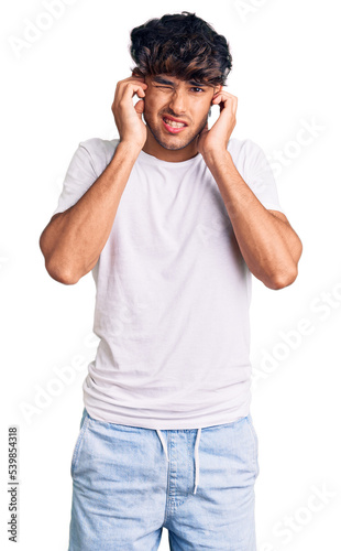 Young hispanic man wearing casual clothes covering ears with fingers with annoyed expression for the noise of loud music. deaf concept.