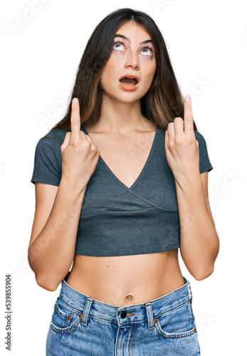 Young beautiful teen girl wearing casual crop top t shirt amazed and surprised looking up and pointing with fingers and raised arms.