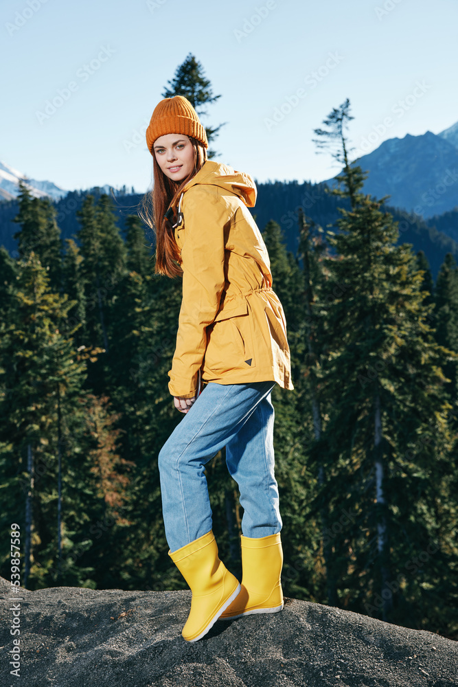 Young woman with red hair full-length hiker in yellow raincoat and cap runs and walks on a mountain trip in the fall and hiking in the mountains at sunset freedom