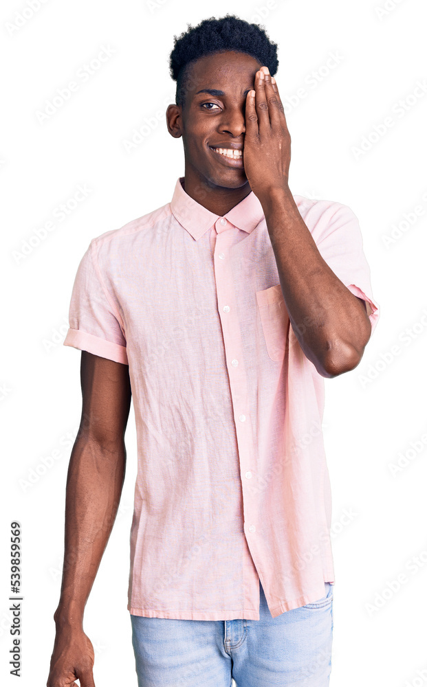 Fototapeta premium Young african american man wearing casual clothes covering one eye with hand, confident smile on face and surprise emotion.