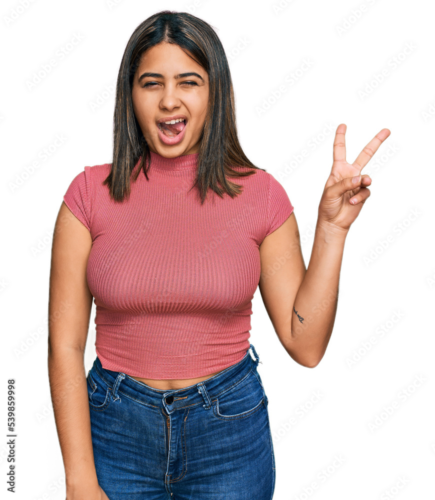 Young hispanic girl wearing casual t shirt smiling with happy face winking at the camera doing victory sign. number two.