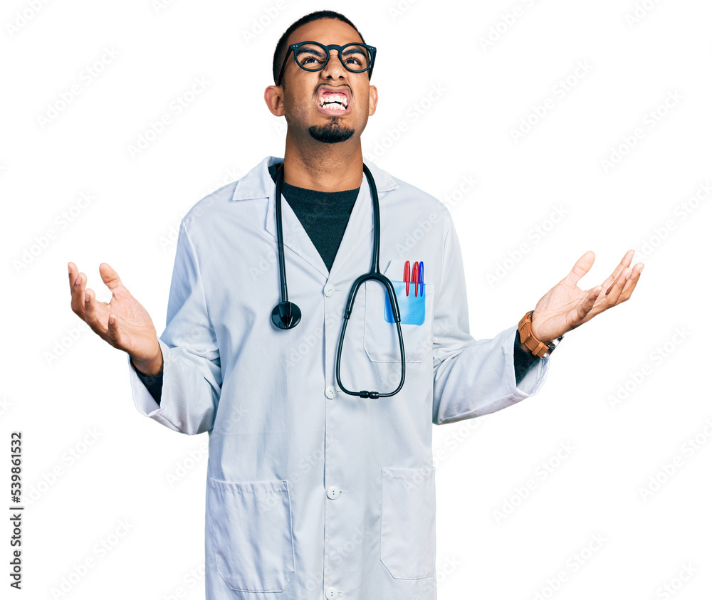 Young african american man wearing doctor uniform and stethoscope crazy and mad shouting and yelling with aggressive expression and arms raised. frustration concept. Young african american man wearing doctor uniform and stethoscope crazy and mad shouting and yelling with aggressive expression and arms raised. frustration concept.