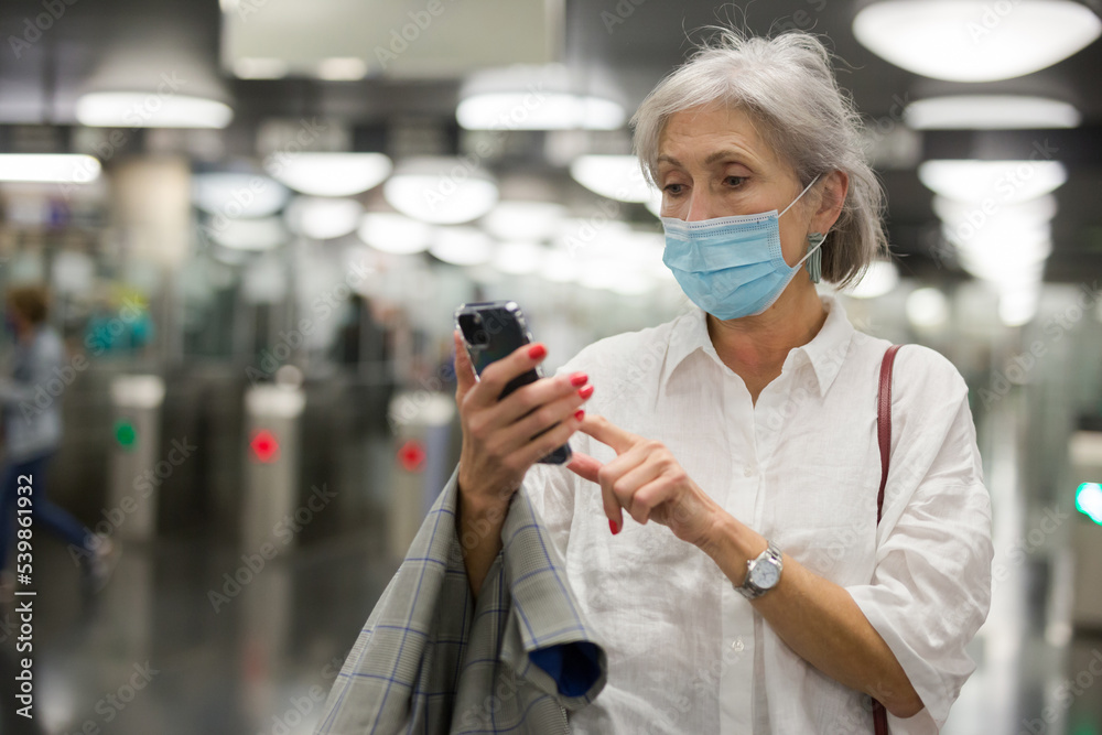 European mature woman in face mask standing near ticket barriers in metro station with her smartphone in hands.