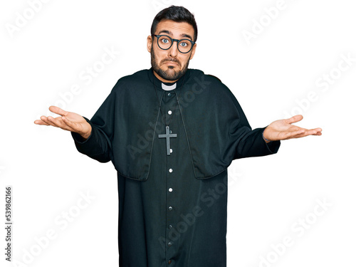 Fotografie, Obraz Young hispanic man wearing priest uniform clueless and confused expression with arms and hands raised