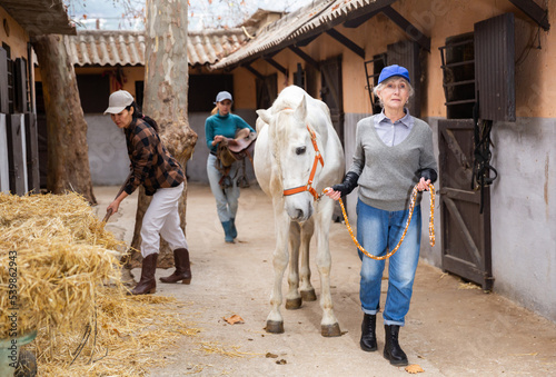 Fotografie Elderly woman horse farm owner leading white racehorse along stalls outdoor while Asian female worker stacking hay with pitchfork in yard