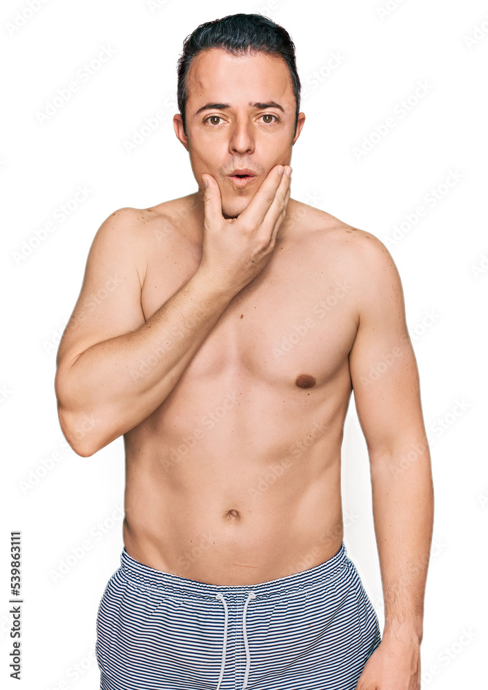 Handsome young man wearing swimwear shirtless looking fascinated with disbelief, surprise and amazed expression with hands on chin Handsome young man wearing swimwear shirtless looking fascinated with disbelief, surprise and amazed expression with hands on chin