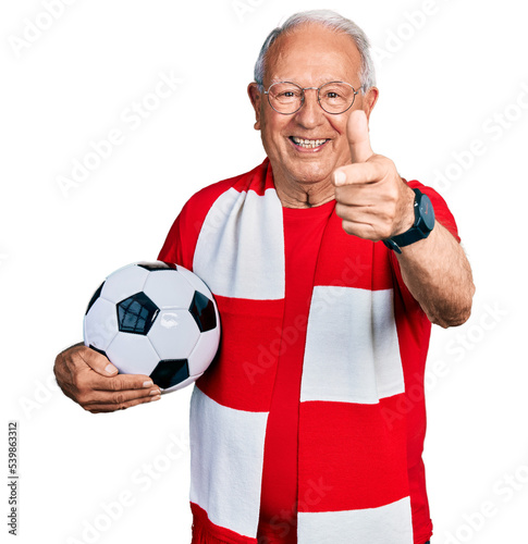 Senior man with grey hair football hooligan holding ball pointing fingers to camera with happy and funny face. good energy and vibes.