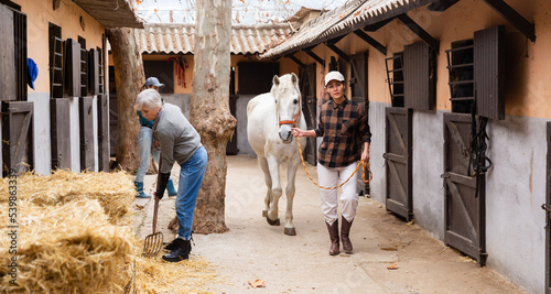 Woman walks a white horse while others clean up the courtyard of the stable