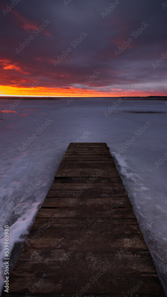 an old boardwalk pier on a large frozen lake with a dramatic evening sky and sunset crimson over the horizon