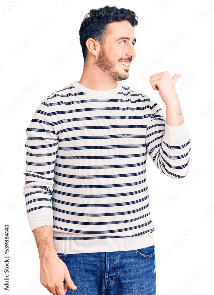 Young hispanic man wearing casual clothes smiling with happy face looking and pointing to the side with thumb up. Young hispanic man wearing casual clothes smiling with happy face looking and pointing to the side with thumb up.