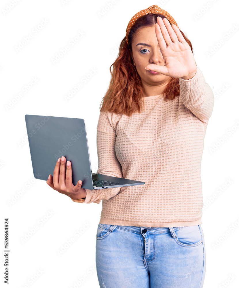 Young latin woman holding laptop with open hand doing stop sign with serious and confident expression, defense gesture Young latin woman holding laptop with open hand doing stop sign with serious and confident expression, defense gesture