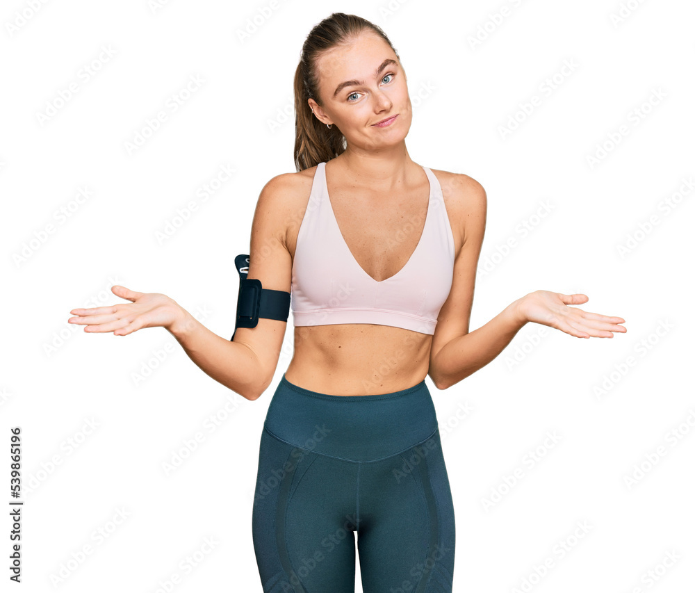 Beautiful young blonde woman wearing sportswear and arm band clueless and confused expression with arms and hands raised. doubt concept. Beautiful young blonde woman wearing sportswear and arm band clueless and confused expression with arms and hands raised. doubt concept.