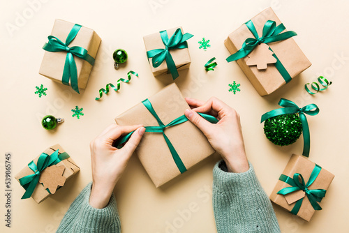 Woman is packing surprise gift for Christmas holidays, sitting at workplace with craft tools, top view. Woman wrapping Christmas gift at colored table