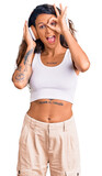 Young hispanic woman with tattoo listening to music using headphones smiling happy doing ok sign with hand on eye looking through fingers