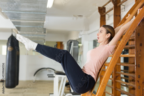 Fit slim young woman training in gym, performing leg raises on wall bars. Bodyweight exercises to strengthen abs muscles. Fitness concept photo