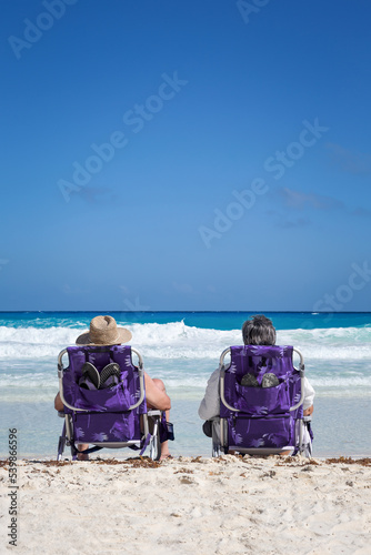 Two unrecognizable people in straw hats sit in chairs on caribbean beach looking to the sea