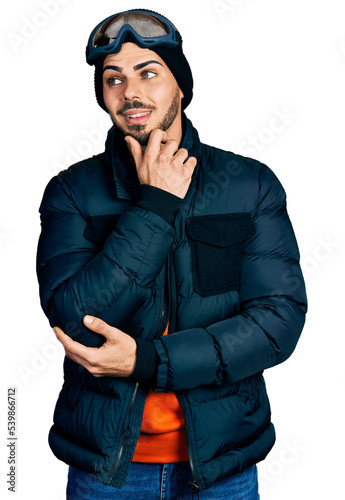 Young hispanic man with beard wearing snow wear and sky glasses with hand on chin thinking about question, pensive expression. smiling with thoughtful face. doubt concept. © Krakenimages.com