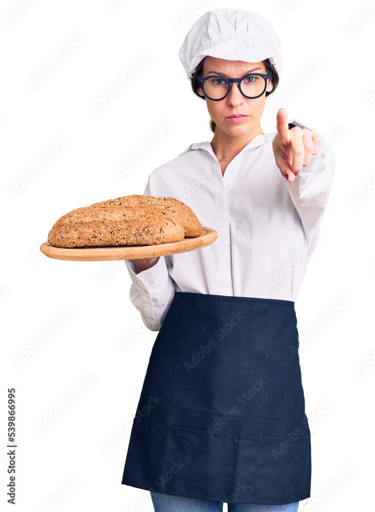 Beautiful brunette young woman wearing baker uniform holding homemade bread pointing with finger to the camera and to you, confident gesture looking serious Beautiful brunette young woman wearing baker uniform holding homemade bread pointing with finger to the camera and to you, confident gesture looking serious