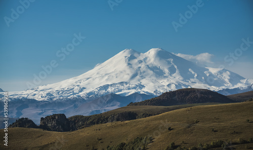 The majestic Mount Elbrus with snow and glaciers and the mountainous Caucasian ridge in the morning light in autumn, with hills with vegetation on a cloudless autumn morning