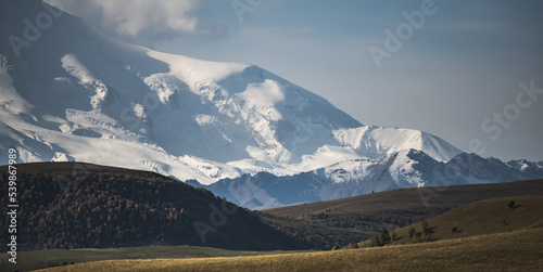 Snowy glacial slope of the Caucasus mountain range and highland hills with trees and shrubs, summer sunny day in the mountains