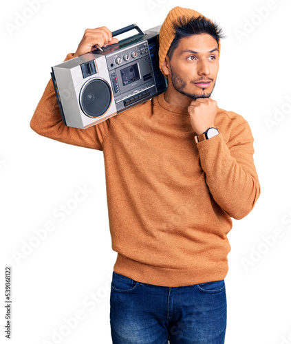 Handsome latin american young man holding boombox, listening to music with hand on chin thinking about question, pensive expression. smiling and thoughtful face. doubt concept.
