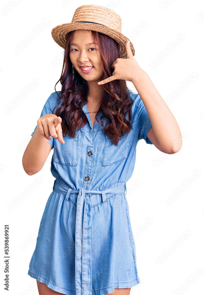 Young beautiful chinese girl wearing summer hat smiling doing talking on the telephone gesture and pointing to you. call me. Young beautiful chinese girl wearing summer hat smiling doing talking on the telephone gesture and pointing to you. call me.