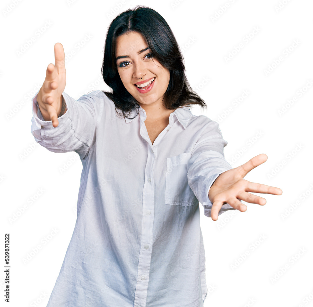 Beautiful hispanic woman wearing casual white shirt looking at the camera smiling with open arms for hug. cheerful expression embracing happiness. Beautiful hispanic woman wearing casual white shirt looking at the camera smiling with open arms for hug. cheerful expression embracing happiness.