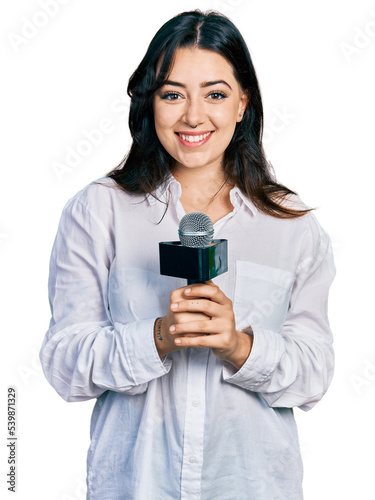 Beautiful hispanic woman holding reporter microphone looking positive and happy standing and smiling with a confident smile showing teeth