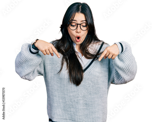 Beautiful hispanic woman wearing casual sweater and glasses pointing down with fingers showing advertisement, surprised face and open mouth