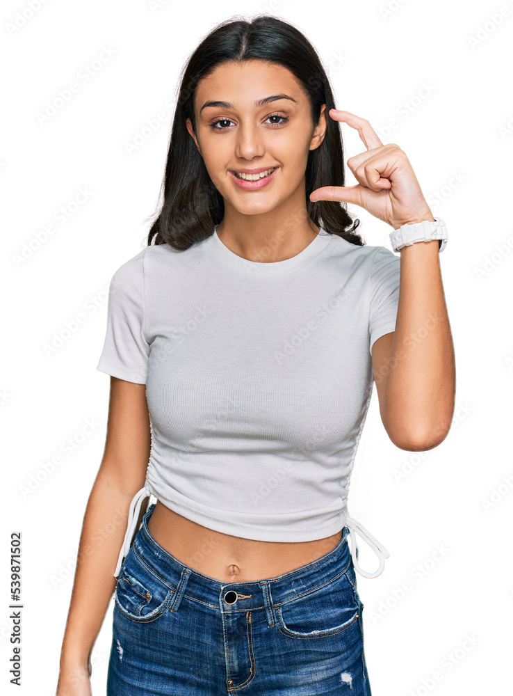 Young hispanic girl wearing casual white t shirt smiling and confident gesturing with hand doing small size sign with fingers looking and the camera. measure concept. Young hispanic girl wearing casual white t shirt smiling and confident gesturing with hand doing small size sign with fingers looking and the camera. measure concept.