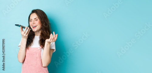 Portrait of smiling woman holding phone near lips and talking, using app translator on smartphone or recording voice message, speaking with speakerphone, standing over blue background