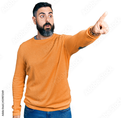 Hispanic man with beard wearing casual winter sweater pointing with finger surprised ahead, open mouth amazed expression, something on the front