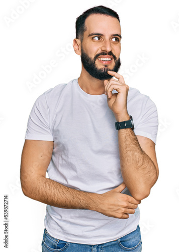 Young man with beard wearing casual white t shirt with hand on chin thinking about question, pensive expression. smiling and thoughtful face. doubt concept.