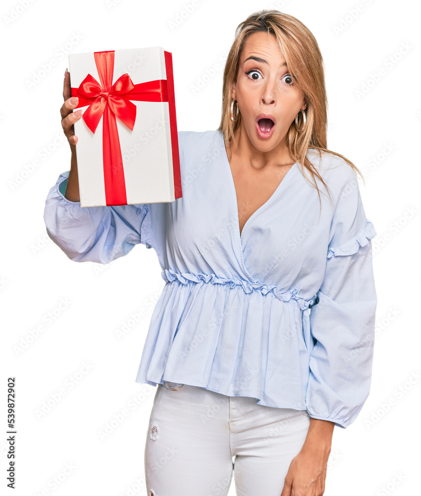 Beautiful blonde woman holding gift scared and amazed with open mouth for surprise, disbelief face Beautiful blonde woman holding gift scared and amazed with open mouth for surprise, disbelief face