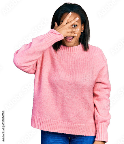 Beautiful hispanic woman wearing casual winter sweater peeking in shock covering face and eyes with hand, looking through fingers with embarrassed expression.